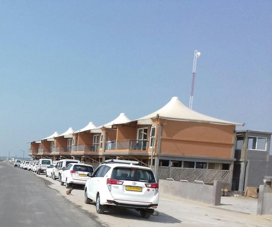A row of white cars parked along a road with a row of buildings featuring balconies and a single tower.