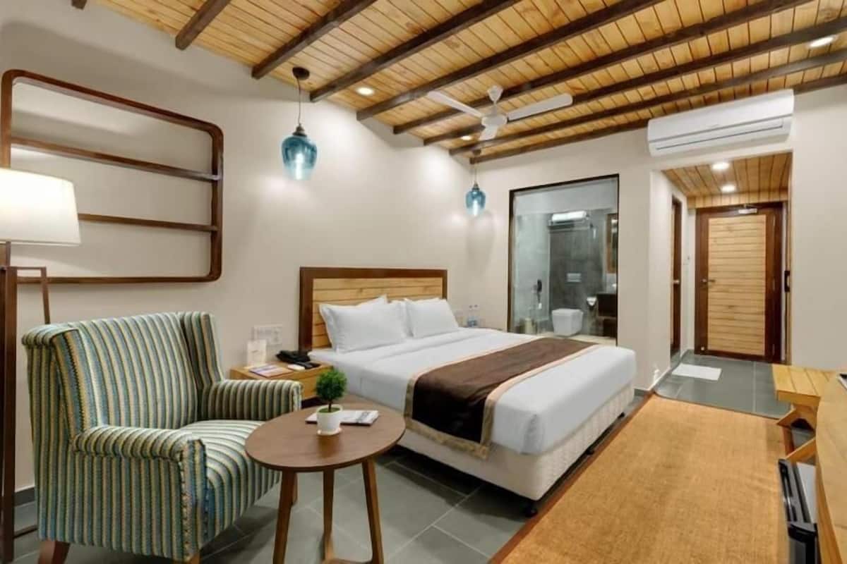 A hotel room with a bed, a chair, a small table, a desk, a TV, and an air conditioning unit.