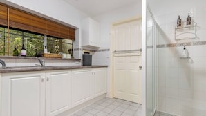 Une cuisine avec des meubles blancs, une fenêtre avec des stores, une porte et un espace douche doté de portes en verre.