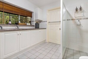 A kitchen with white cabinets, a window with blinds, a door, and a shower area with glass doors.