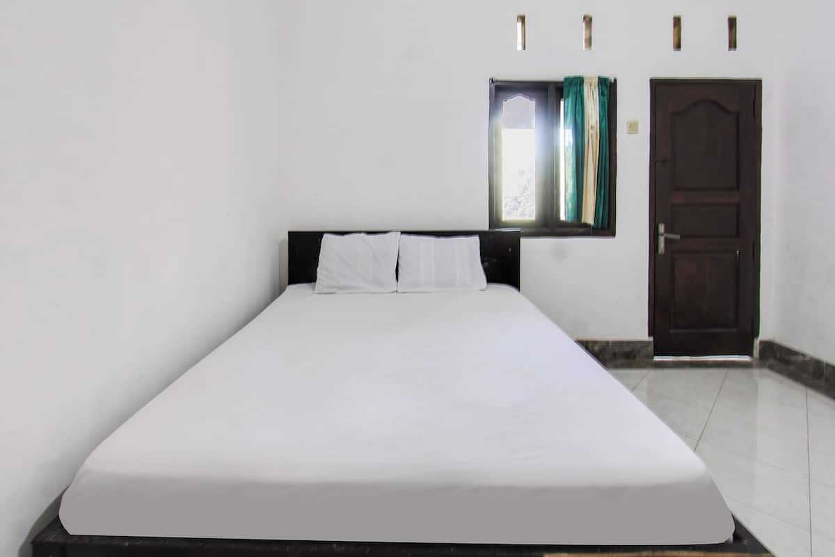 Uma cama de solteiro com roupa de cama branca em um quarto com paredes brancas, uma porta de madeira e uma janela com cortinas.