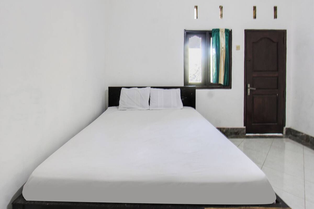 Un lit simple avec une literie blanche, dans une pièce aux murs blancs, avec une porte en bois et une fenêtre ornée de rideaux.
