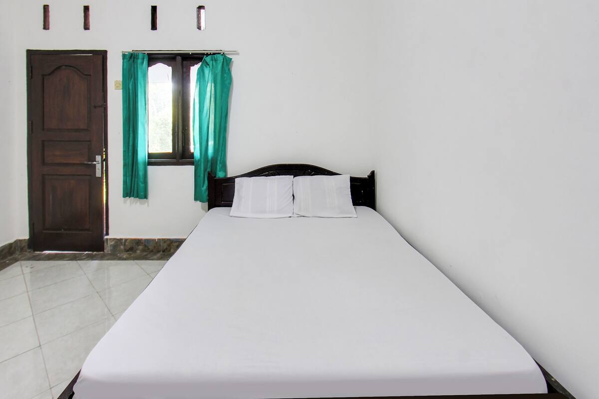 Un lit simple avec du linge de lit blanc, une tête de lit en bois et une fenêtre ornée de rideaux verts.