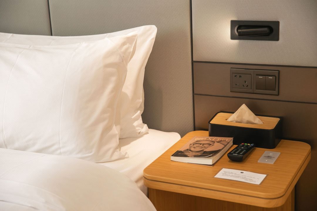 A hotel room with a bed, a bedside table with a book, a remote control, a tissue box, and a power outlet.