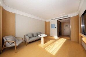 Una habitación de hotel moderna con piso de madera, un sofá gris, una silla de madera, una mesita redonda y un televisor.
