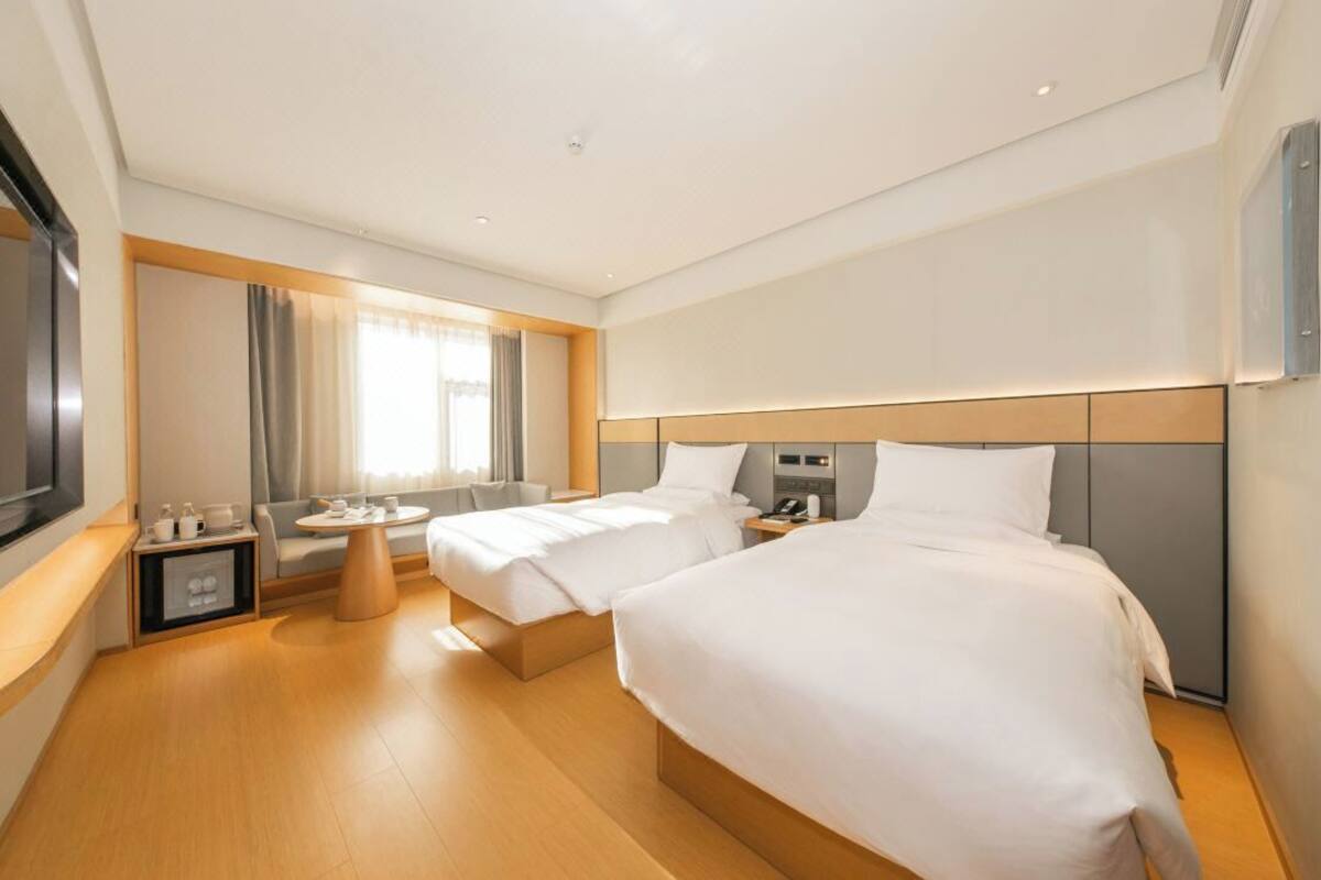 A hotel room with two beds, a bedside table, a small round table, a TV, and a window with curtains.
