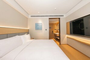 Quarto de hotel com cama, janela grande, espelho e porta para o banheiro.