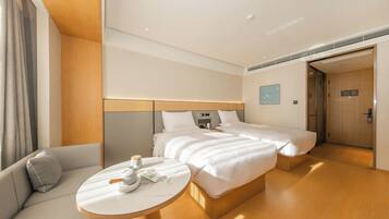 A hotel room with a sofa, a round table, two beds, a wooden headboard, and a door leading to another room.