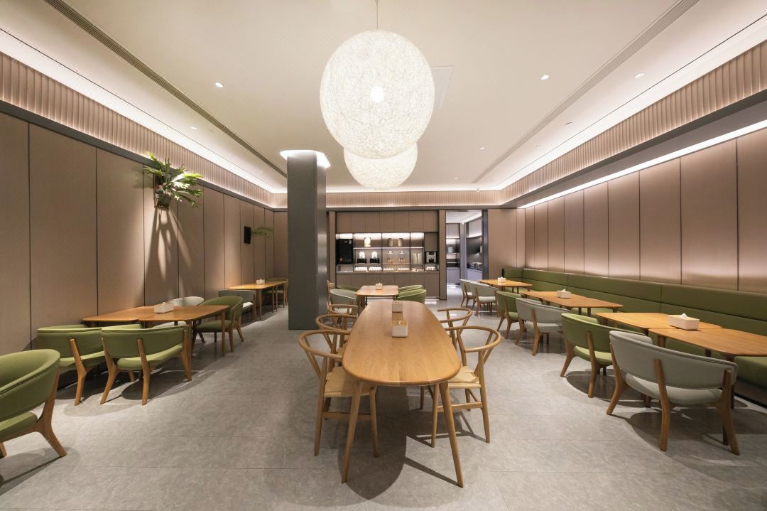 Un comedor moderno con mesas y sillas de madera, una lámpara central y una cocina al fondo.