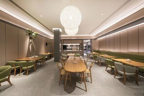 Un comedor moderno con mesas y sillas de madera, una lámpara central y una cocina al fondo.