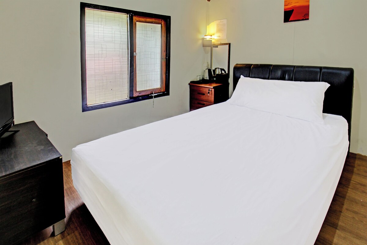 A neatly made bed with a white sheet, a wooden nightstand with a lamp, a small table with a cup, and a window with blinds.