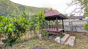 Um gazebo de madeira com telhado de azulejos, rodeado por vegetação exuberante e uma encosta ao fundo.