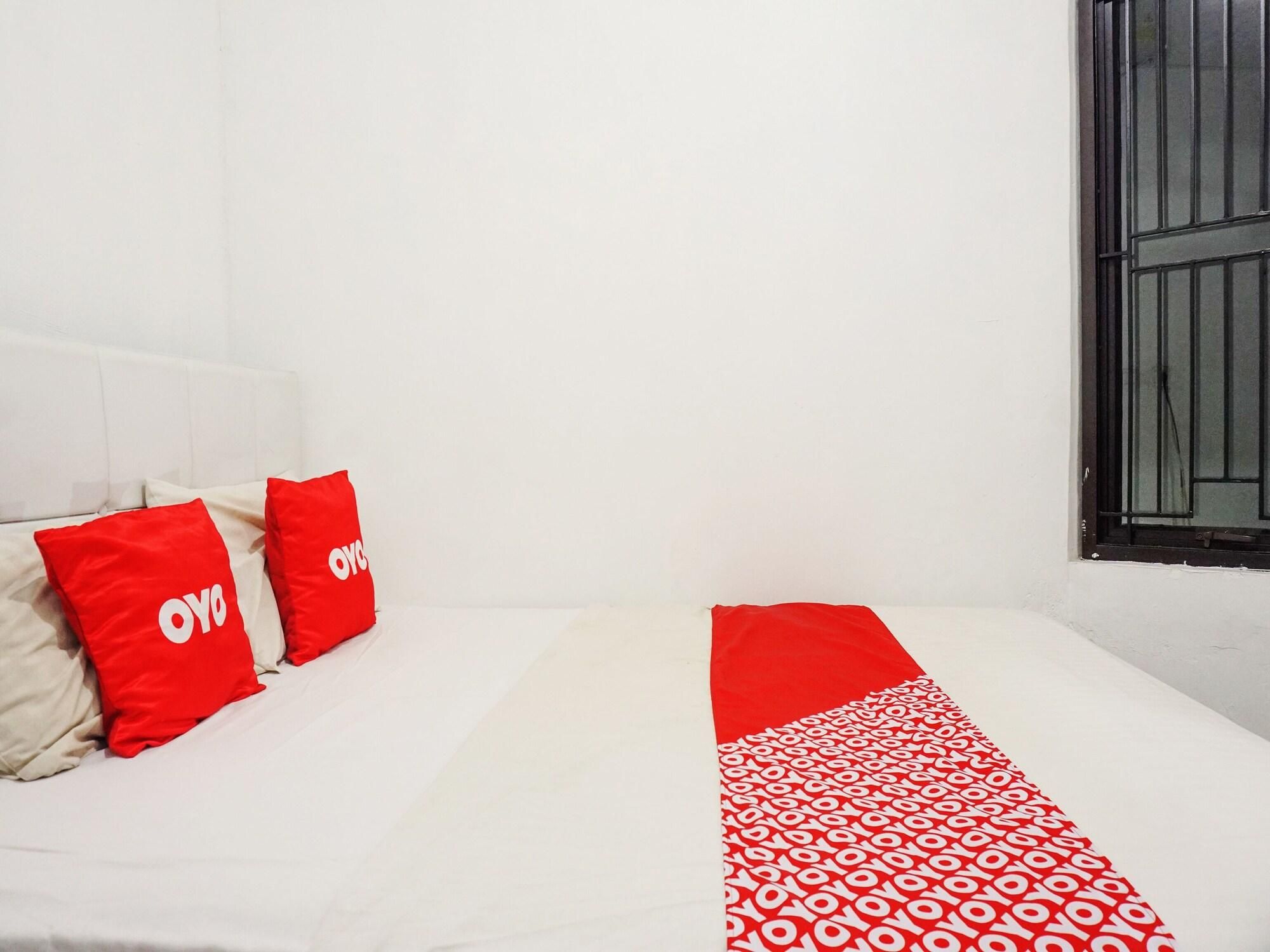 Una cama con ropa de cama blanca y detalles en rojo, una ventana con barrotes negros y una pared blanca.