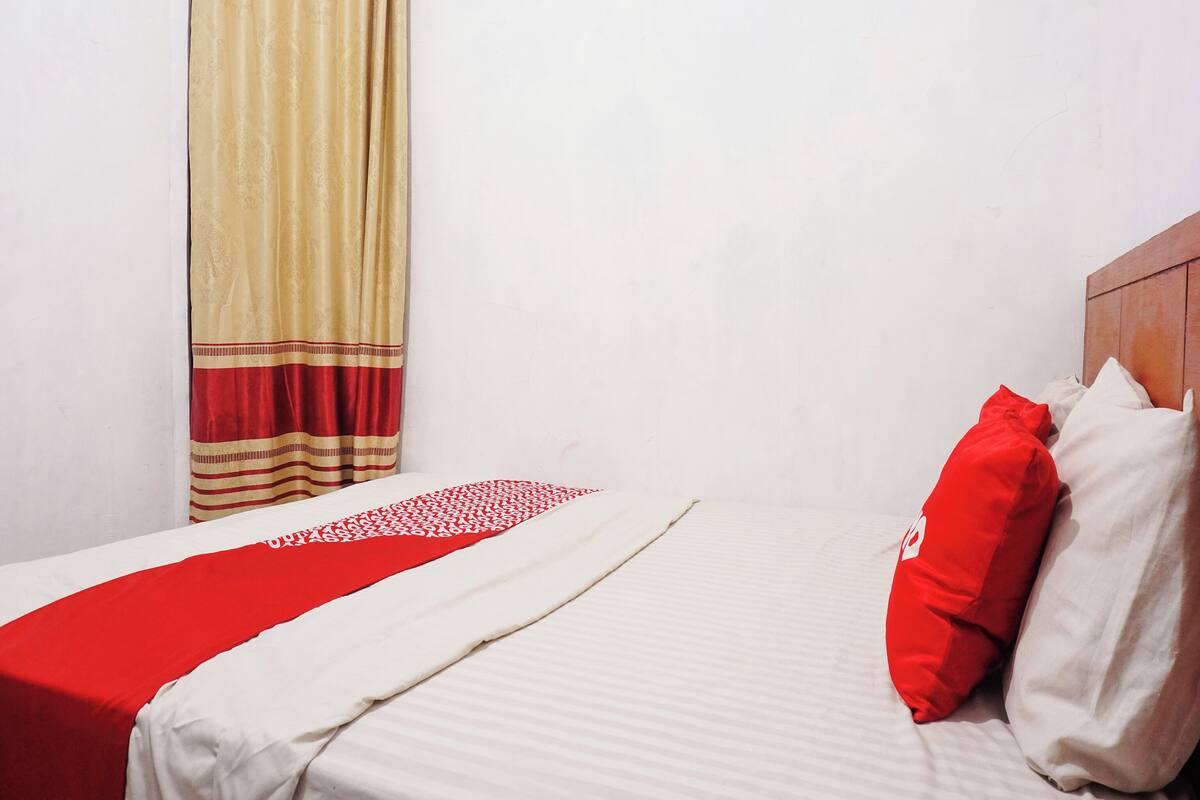 A neatly made bed with a red and white bedspread, a wooden headboard, and a window with beige curtains.