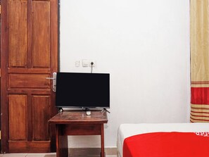 Una habitació amb una porta de fusta, un televisor damunt d'una taula de fusta petita, un llit amb col·cha vermella i blanca, i una paret blanca sense adorns amb una presa elèctrica.