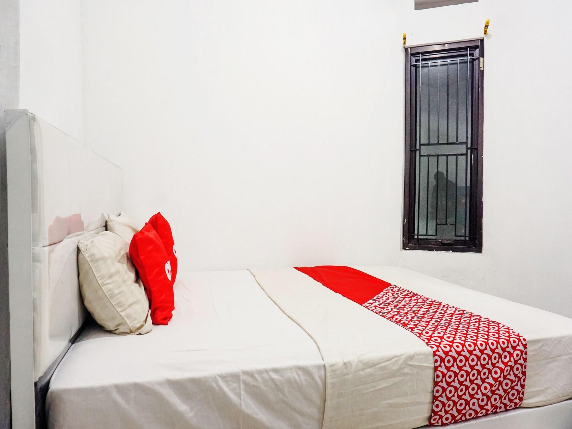 A single bed with a white and red bedspread, a red pillow, and a window with a metal grill.