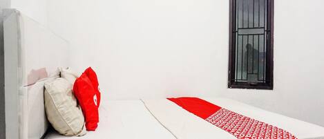A single bed with a white and red bedspread, a red pillow, and a window with a metal grill.