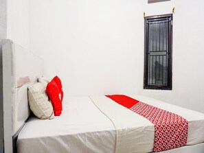 Una cama individual con una colcha blanca y roja, un almohón rojo y una ventana con reja de metal.