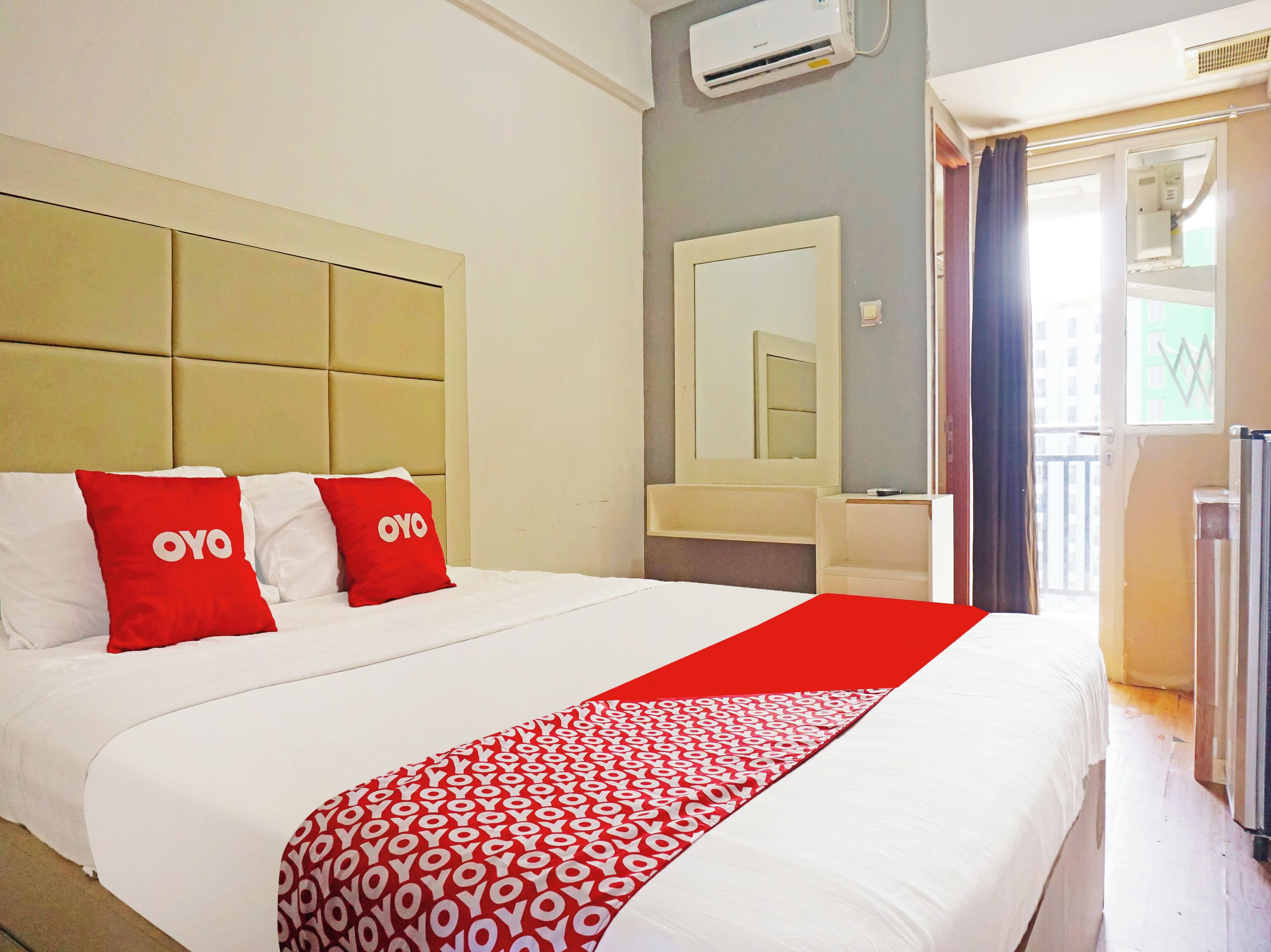 A hotel room with a large bed, two red pillows with 'OYO' branding, a white bedspread, a red and white patterned throw, a large mirror, and a window with curtains.