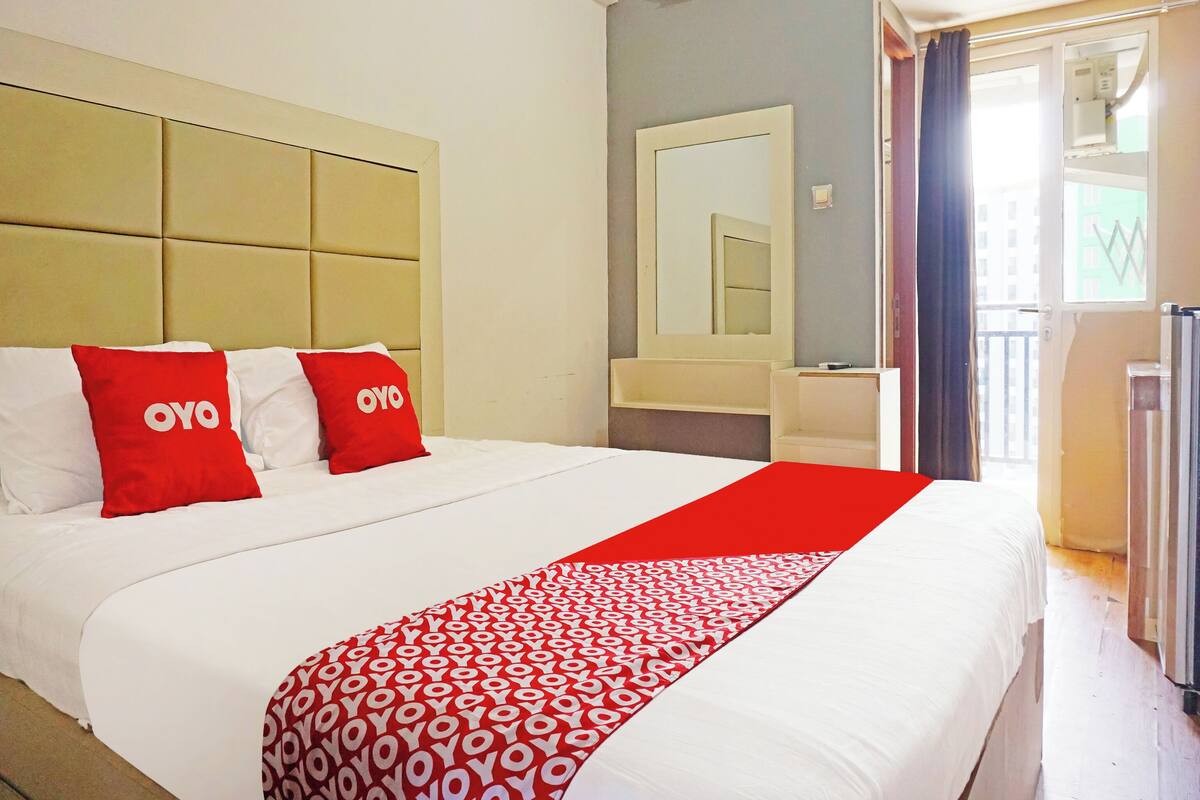 Habitación de hotel con una cama grande, dos almohadas rojas con la marca 'OYO', una colcha blanca, un plaid rojo y blanco estampado, un espejo grande y una ventana con cortinas.