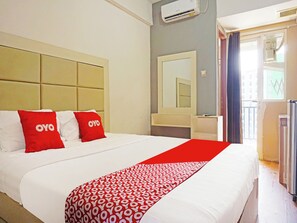 Quarto de hotel com uma cama grande, duas almofadas vermelhas com a marca 'OYO', uma colcha branca, um cobertor estampado em vermelho e branco, um espelho grande e uma janela com cortinas.