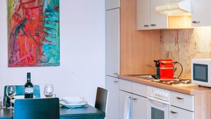 Ett kompakt kök med vita skåp, en mikrovågsugn, en röd vattenkokare och ett matbord med stolar.