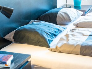 Un letto rifatto con biancheria azzurra e bianca, un comodino con una lampada e un libro sul tavolo.