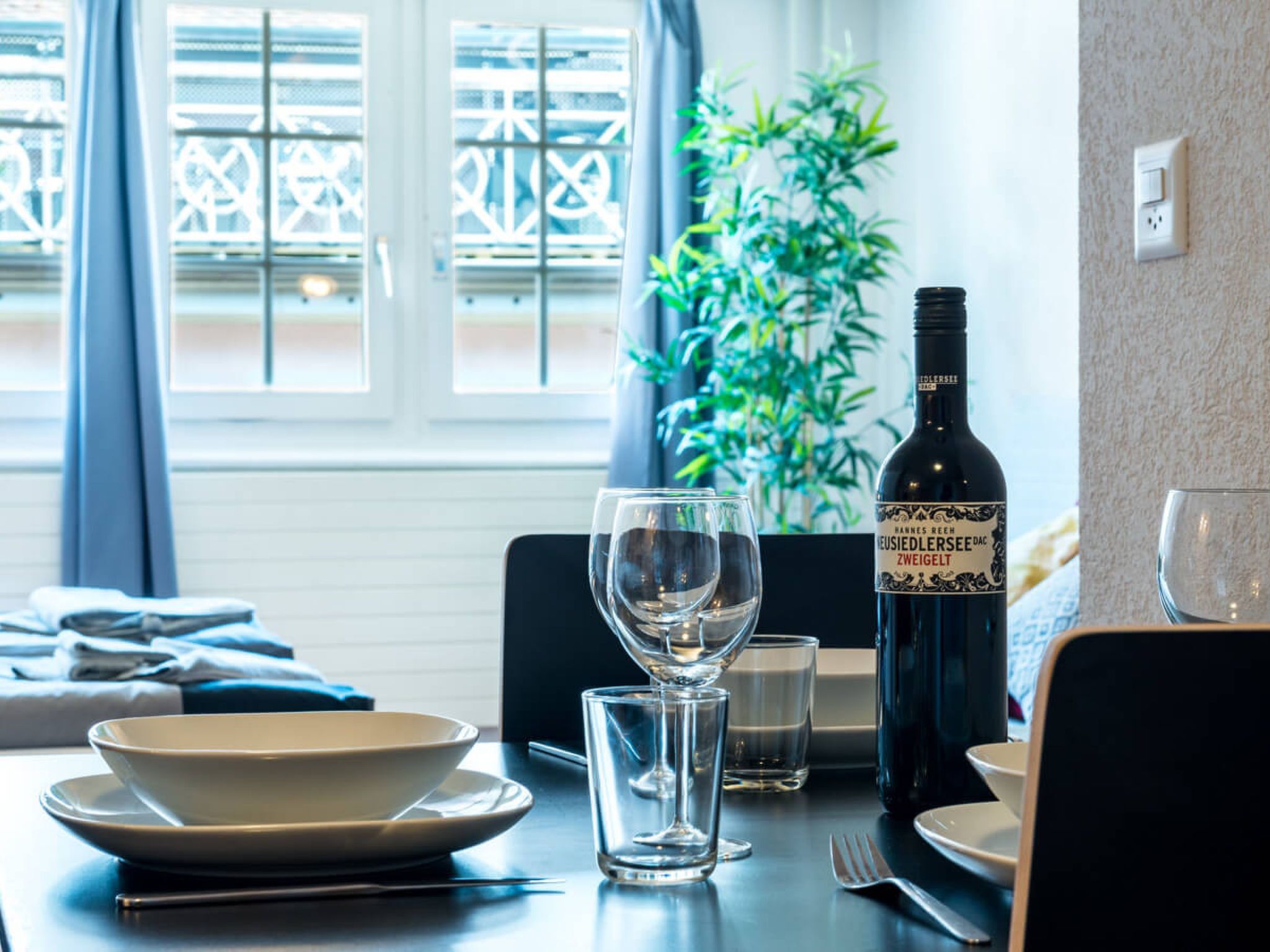 A dining table with a bottle of wine, a glass, and plates, set against a window with a view of a building.