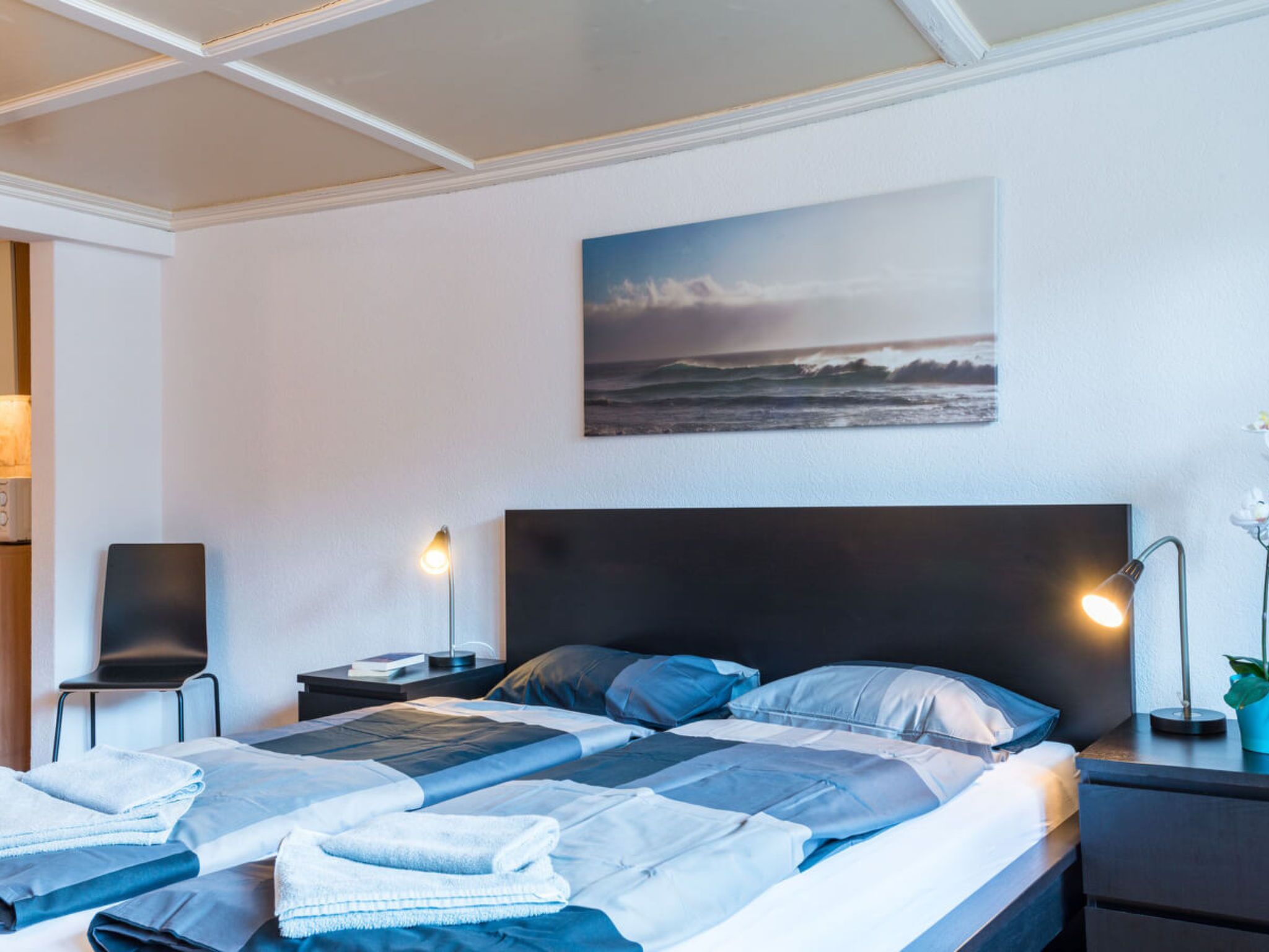 Une chambre d’hôtel avec un lit, deux tables de chevet, une petite table, un tableau accroché au mur et une plante en pot.