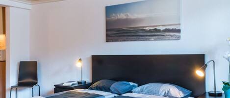 Une chambre d’hôtel avec un lit, deux tables de chevet, une petite table, un tableau accroché au mur et une plante en pot.