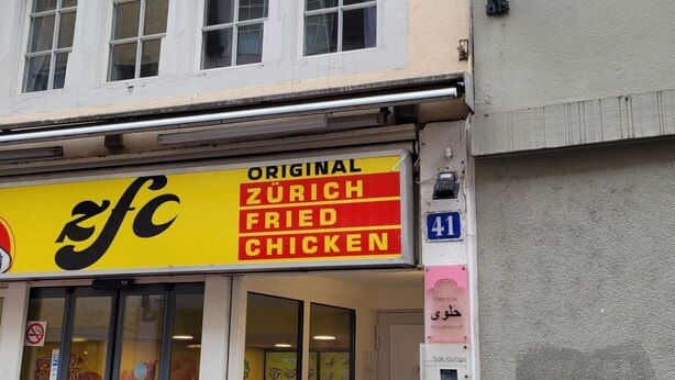 En byggnad med en gul skylt som visar namnet på en restaurang: ”ZFC Original Zurich Fried Chicken”.