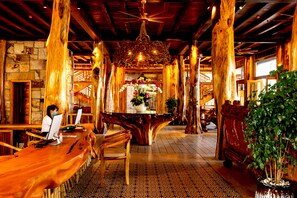 A rustic interior with wooden beams, a central wooden table with a vase of flowers, and a person using a laptop at a long wooden table. - Ocean Bay Resort & Spa Phu Quoc (Phu Quoc)