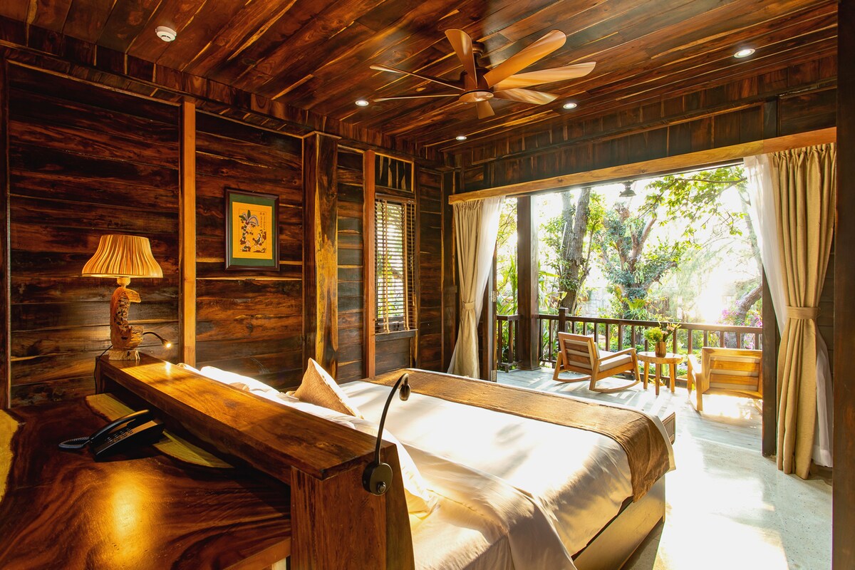 A wooden cabin-style room with a bed, a desk, a chair, and a balcony with a view of trees.
