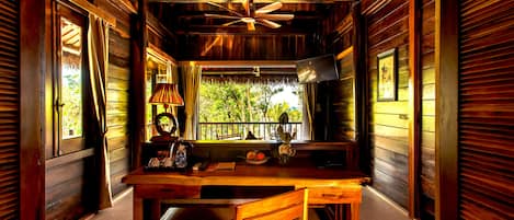 A wooden interior with a table, chairs, a ceiling fan, a window with curtains, and a framed picture on the wall.