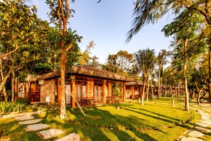 A rustic wooden house with a thatched roof, surrounded by a well-maintained lawn and tall trees. - Ocean Bay Resort & Spa Phu Quoc (Phu Quoc)