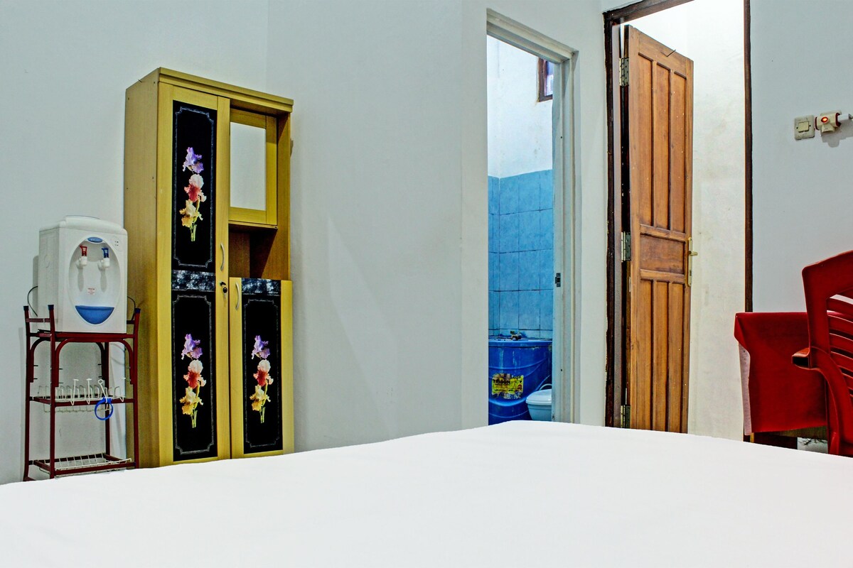 Una habitación con una cama blanca, un armario de madera con decoraciones florales, una silla roja y un dispensador de agua.