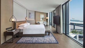 Una habitación de hotel moderna con una cama grande, mesitas de noche, un televisor y vistas al aeropuerto.
