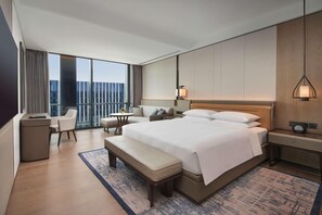 Ein Hotelzimmer mit einem großen Bett, einem Schreibtisch, einem Sessel, einem Nachttisch, einer Lampe und Blick auf die Stadt durch das Fenster.