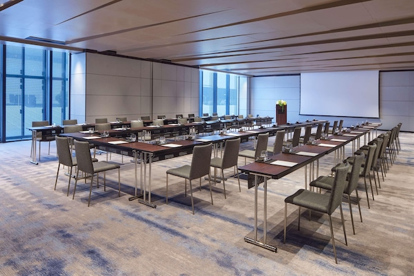 Un salón de conferencias con mesas largas, sillas, una pantalla de proyector y ventanas amplias.