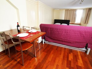Ein kleines Zimmer mit Esstisch, Stühlen, einem Bett mit rosa Bettwäsche und einem Fenster mit Vorhängen.
