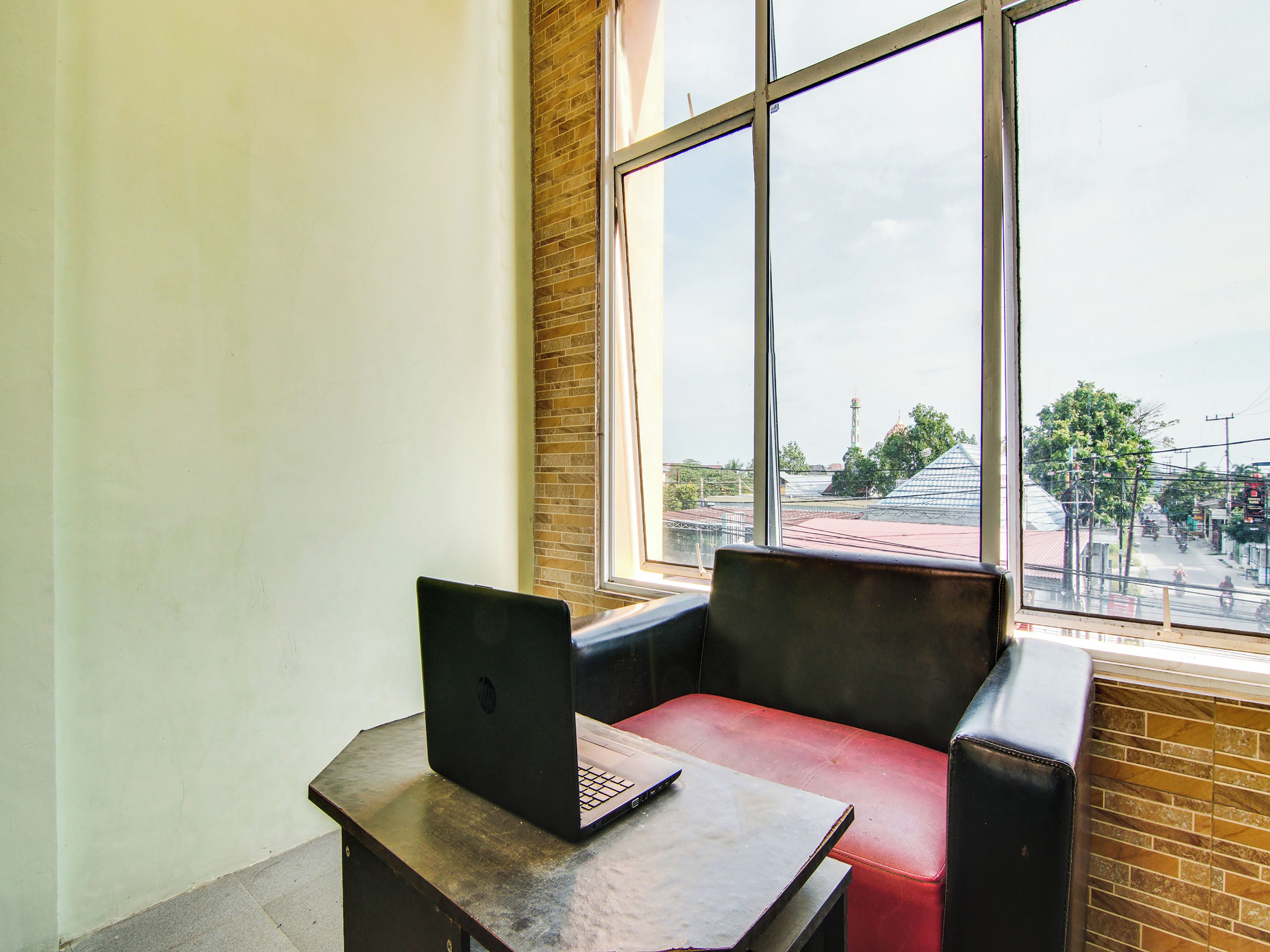 A room with a laptop on a table, a red chair, and a large window offering a view of the street and buildings outside.