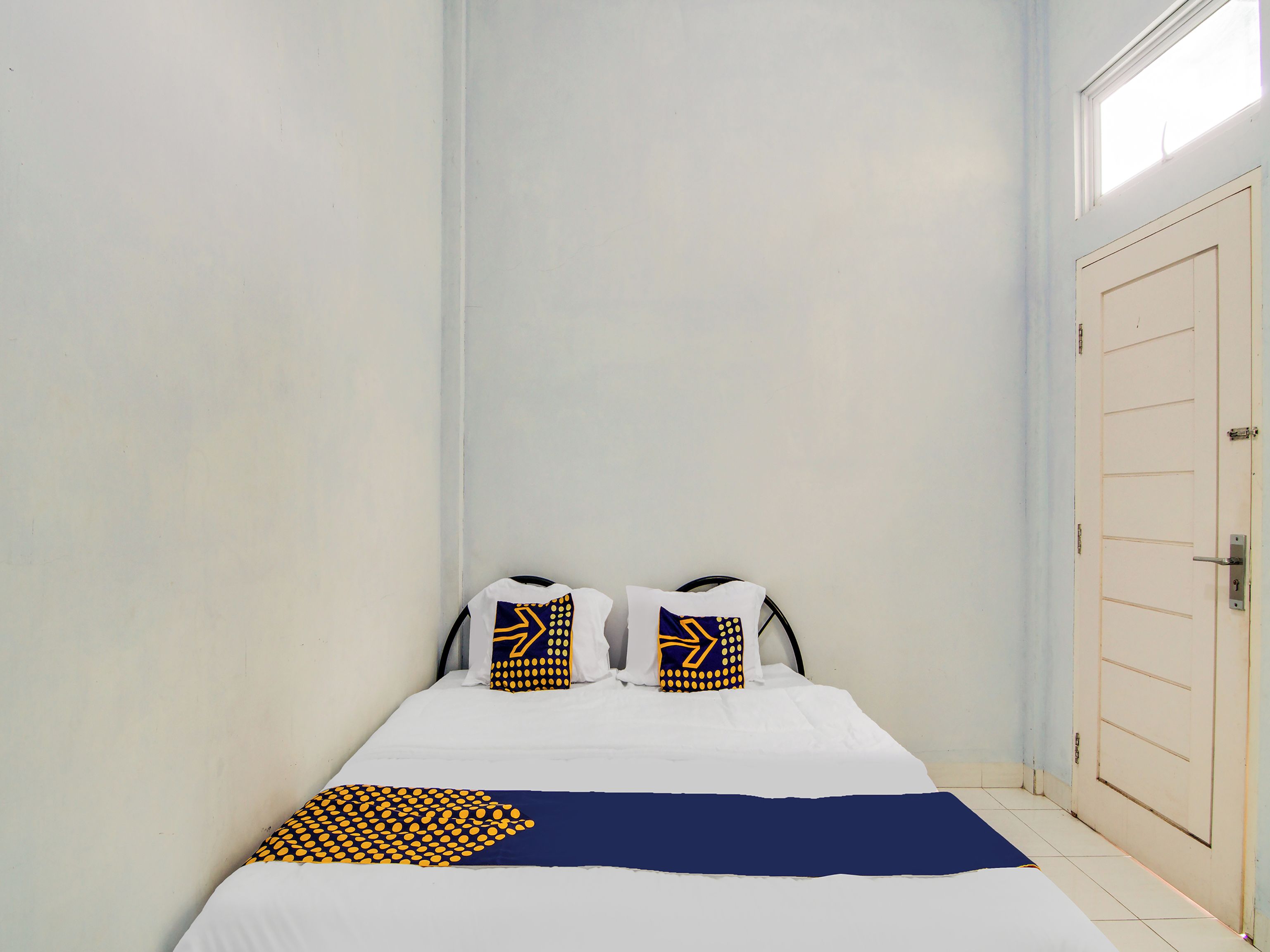 A small, narrow hotel room with a single bed, two pillows with geometric patterns, and a plain white door.