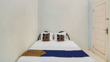 A small, narrow hotel room with a single bed, two pillows with geometric patterns, and a plain white door.