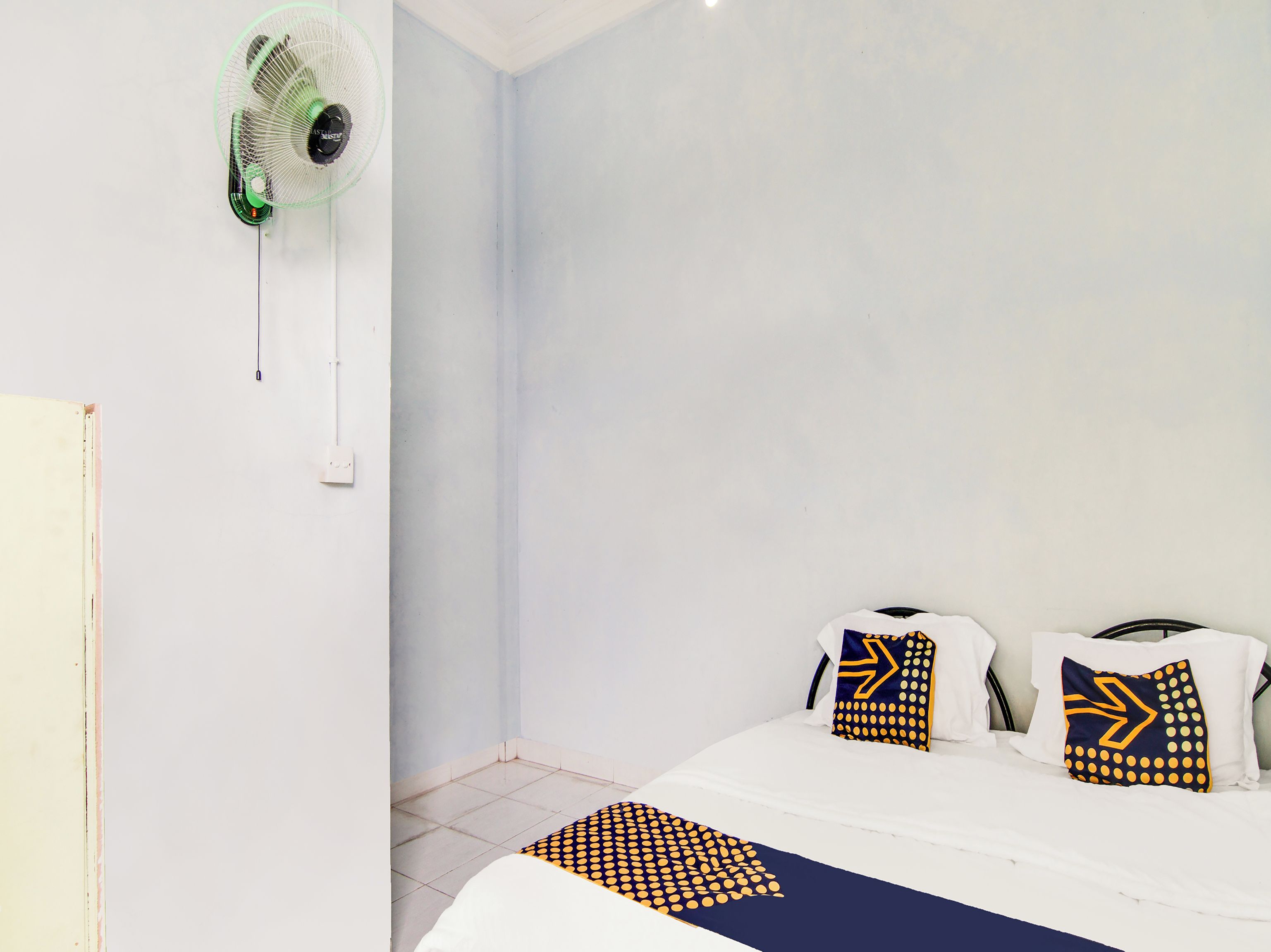 Ein Schlafzimmer mit einem Bett, zwei Kissen mit geometrischen Mustern, einem Standventilator und einer weißen Wand.