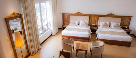 Et hotelværelse med to senge, et opholdsområde med et bord og stole, et spejl og et vindue med gardiner.