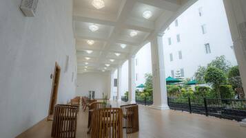 A spacious indoor area with a high ceiling, wooden furniture, and a view of a building with columns and greenery outside.