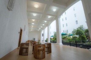 A spacious indoor area with a high ceiling, wooden furniture, and a view of a building with columns and greenery outside. - Araliya Red (Nuwara Eliya)