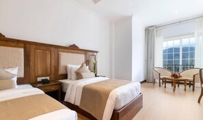 A hotel room with two beds, a wooden headboard, a dining table with chairs, and a large window with curtains. - Araliya Red (Nuwara Eliya)