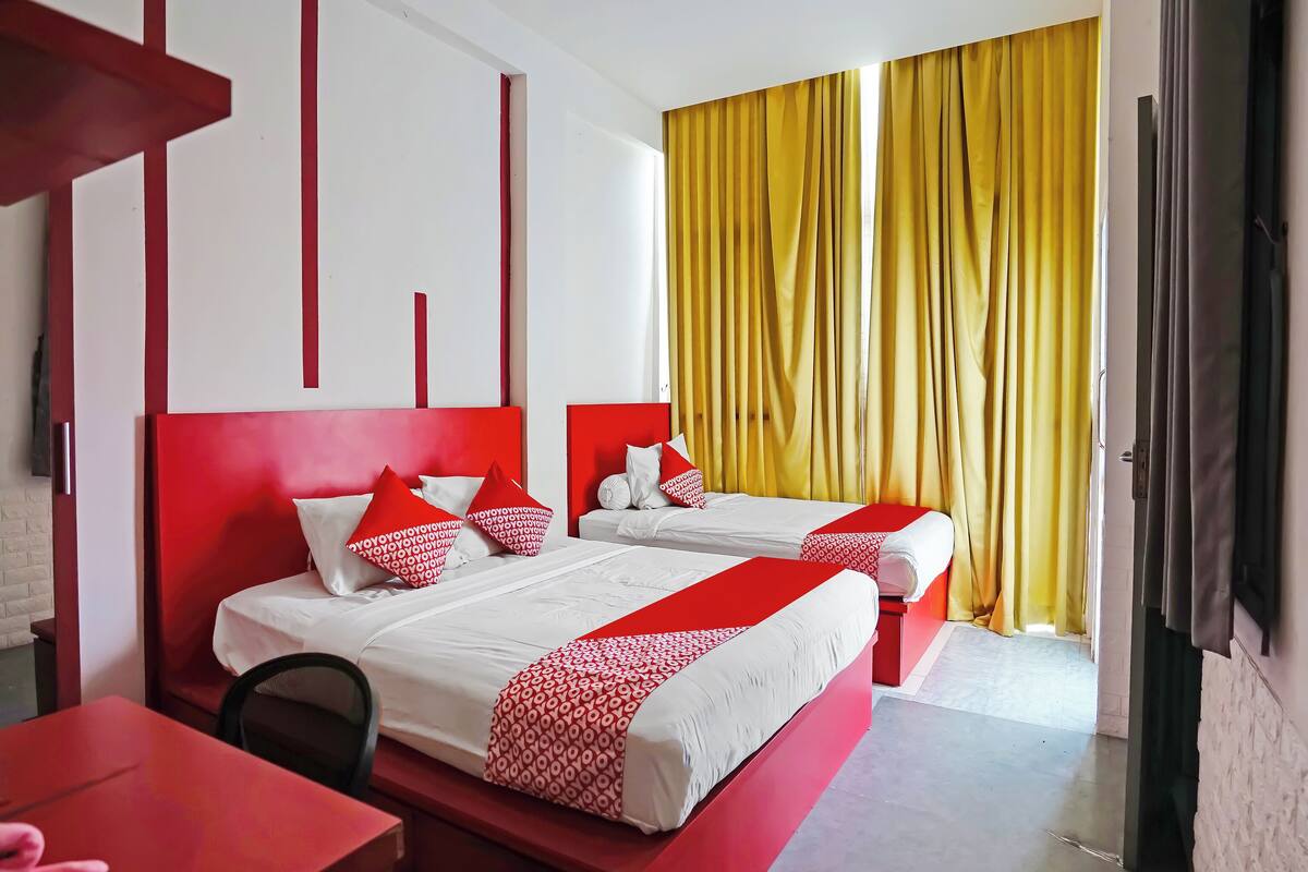 Een hotelkamer met twee bedden, rood-wit beddengoed, een rode tafel en gele gordijnen.