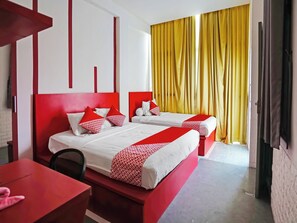 Habitación de hotel con dos camas, ropa de cama roja y blanca, una mesa roja y cortinas amarillas.